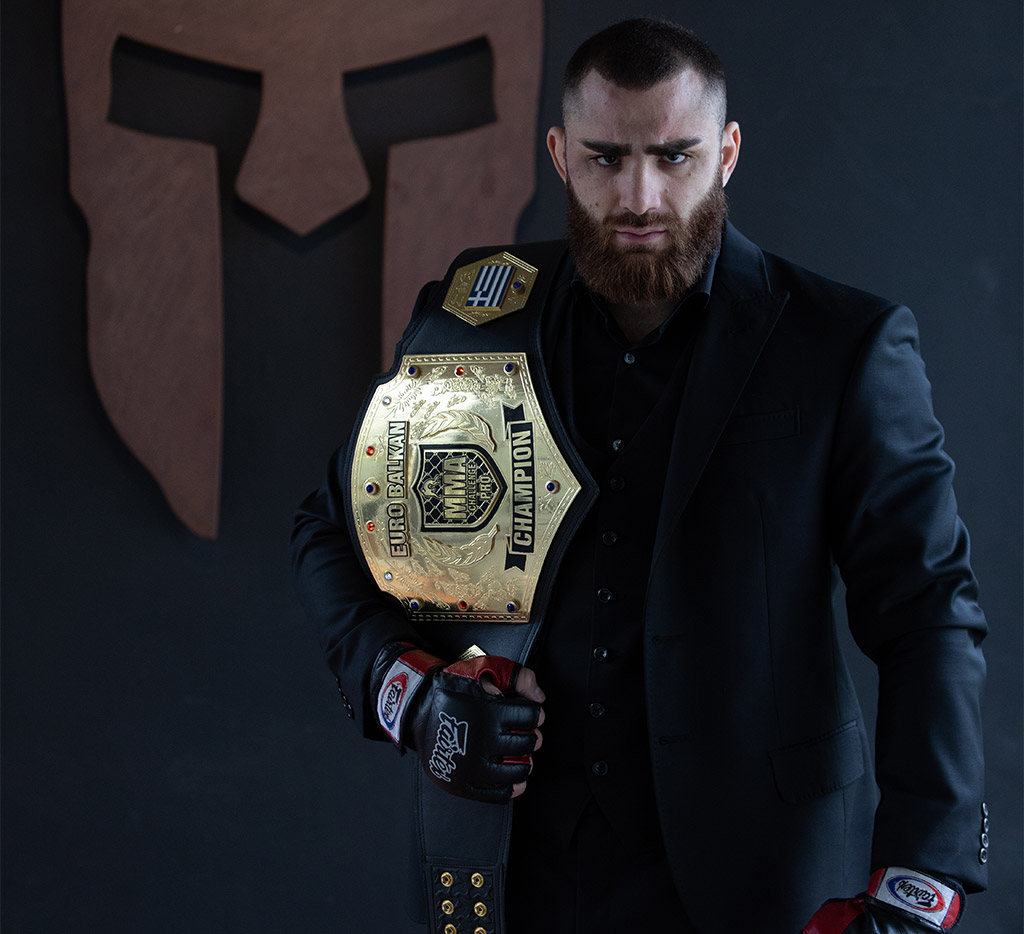Andreas Tricomitis in a black suit holding the EuroBalkan MMA Championship belt
